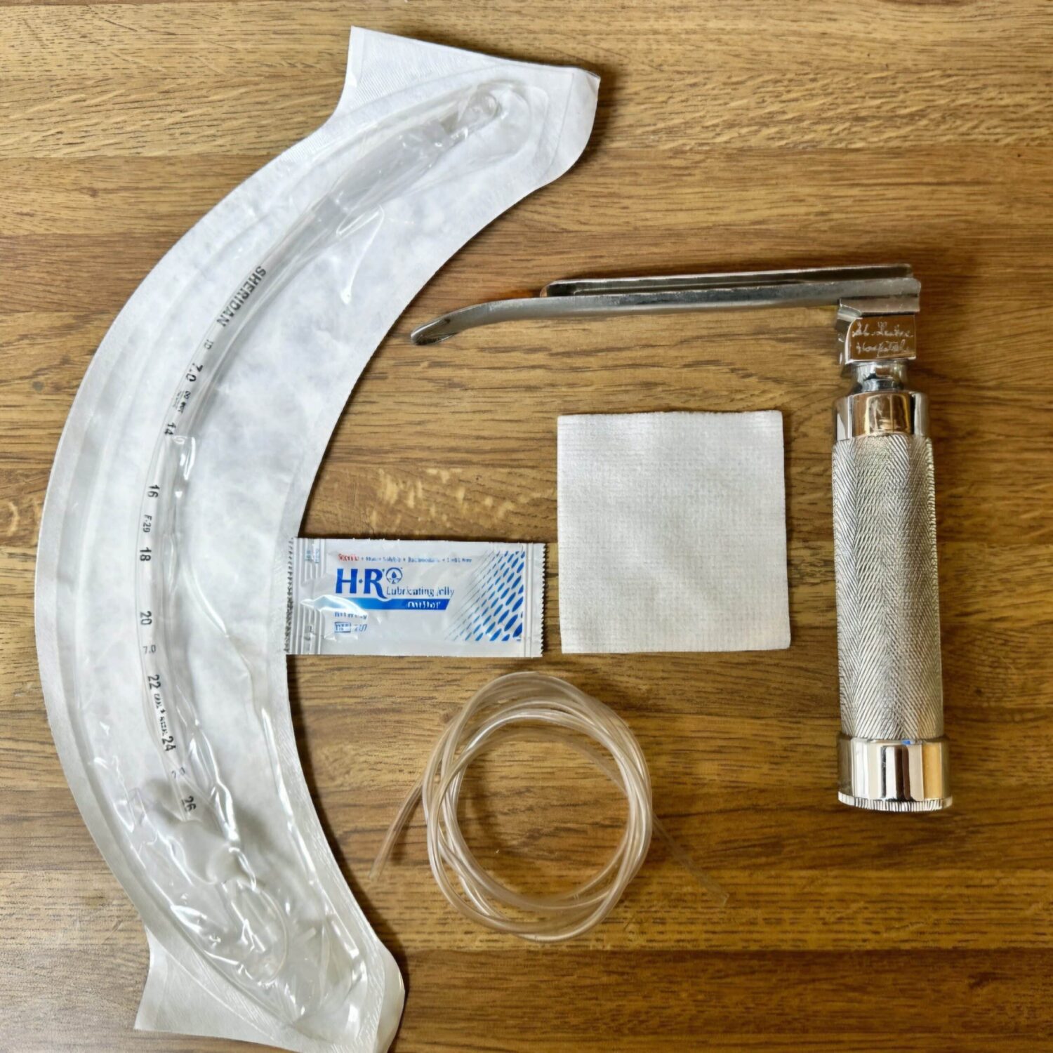 Medical equipment including a sealed endotracheal tube, a laryngoscope, a pack of lubricant, a white gauze, and a clear tube, arranged on a wooden surface.