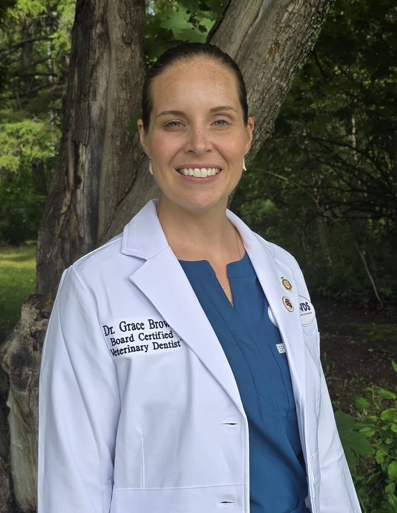 A woman in a white lab coat stands outdoors in front of trees, smiling. The coat reads "Dr. Grace Brown, Board Certified Veterinary Dentist.