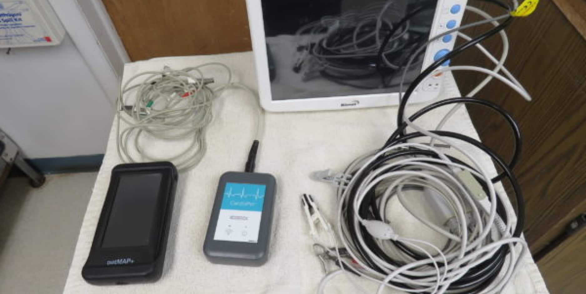 A collection of medical devices and cables are laid out on a white surface, including a monitor, a CenTrak device, a Dex-P portable screen, and various cords and electrodes.