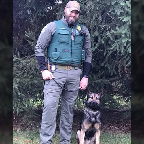 A person in uniform stands outdoors holding a leash connected to a seated German Shepherd dog.