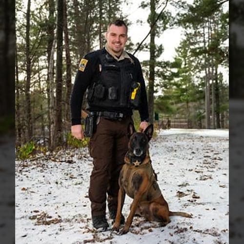 A uniformed officer stands in a snowy wooded area with a police dog.